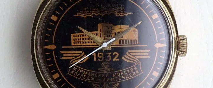 Primo piano di un orologio con quadrante nero e dettagli dorati, recante la scritta in cirillico "1932" e "МУРМАНСКИЙ МОРСКОЙ КОЛЛЕДЖ ИМ. И. МЕСЯЦЕВА". Proprietà di Sovietaly.